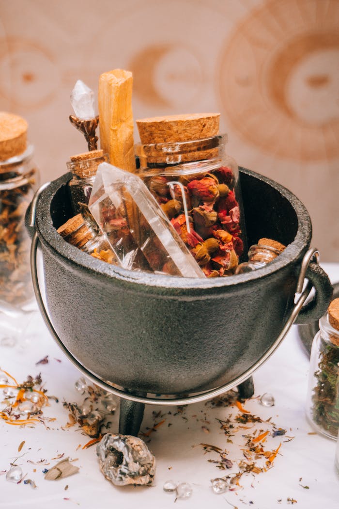 A selection of herbal ingredients and crystals in a rustic cauldron surrounded by jars and dried flowers.
